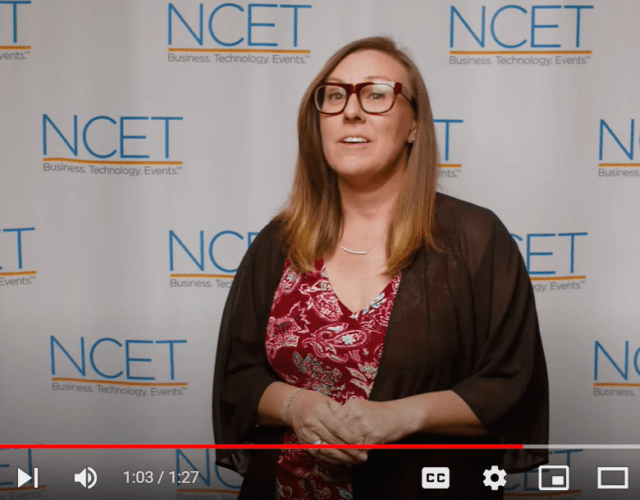 Amy is a caucasian woman with glasses and medium length golden brown hair. She is wearing a black sweater over a red patterned blouse and is speaking in front of the NCET print background wall.