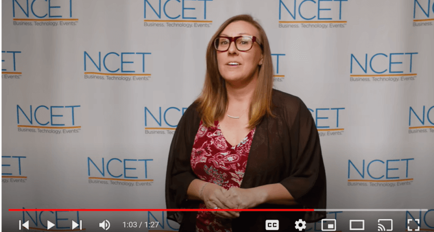 Amy is a caucasian woman with glasses and medium length golden brown hair. She is wearing a black sweater over a red patterned blouse and is speaking in front of the NCET print background wall.