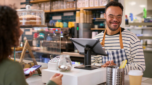 A man with IDD is standing behind a cash register smiling.